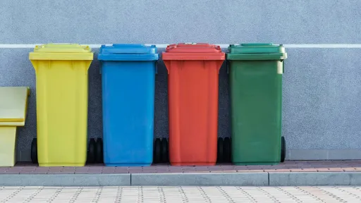 Quatre bacs de collecte des déchets alignés contre un mur gris : un jaune pour les emballages recyclables, un bleu pour le papier, un rouge pour les déchets spécifiques, et un vert pour les biodéchets ou les déchets verts.