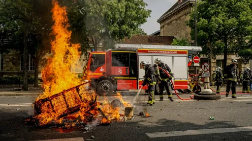 Des sapeurs-pompiers en tenue interviennent pour éteindre un incendie urbain. Un canapé en flammes se trouve au premier plan, tandis qu'un camion de pompiers est stationné en arrière-plan, entouré d'arbres et de bâtiments.