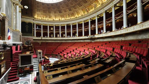 Vue intérieure de l'hémicycle de l'Assemblée nationale française, avec ses sièges rouges disposés en demi-cercle, des colonnes blanches et un plafond orné de décorations dorées. L'espace est vide, sans présence de députés.