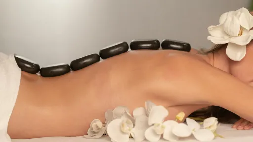 Une femme allongée sur le ventre reçoit un massage aux pierres chaudes. Des pierres noires lisses sont disposées le long de sa colonne vertébrale. Elle porte une fleur blanche dans les cheveux et est entourée de fleurs blanches, créant une ambiance relaxante et apaisante.