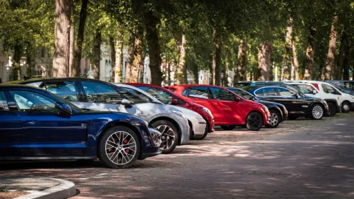 Une rangée de voitures stationnées sur un parking bordé d'arbres, avec des ombres projetées par la lumière du soleil à travers les feuilles.