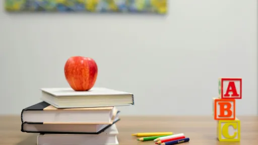 Image d'une pomme rouge posée sur une pile de livres, accompagnée de blocs colorés avec les lettres A, B, C et de crayons de couleur sur une table en bois.