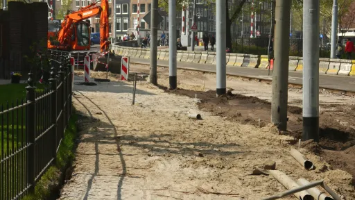 Photo de travaux sur une voie publique, avec une pelleteuse orange, des barrières de sécurité, des panneaux de signalisation, et des éléments de chantier comme des tuyaux et du sable.