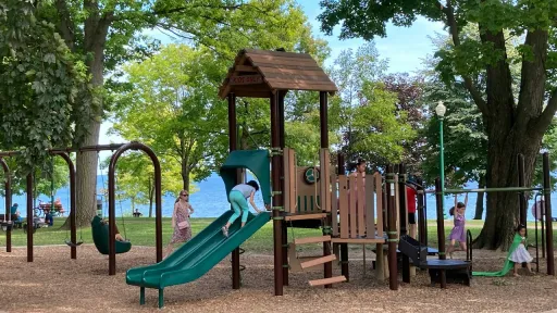 Une aire de jeux en bois avec un toboggan vert et des balan&ccedil;oires, situ&eacute;e dans un parc arbor&eacute; au bord d'un grand lac bleu. Plusieurs enfants jouent sur les structures par une journ&eacute;e ensoleill&eacute;e.