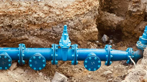 Vue rapproch&eacute;e d'un r&eacute;seau de tuyaux en fonte bleu vif install&eacute;s dans une excavation de terre. On distingue une vanne de sectionnement centrale et plusieurs raccords &agrave; brides fix&eacute;s par des boulons argent&eacute;s, typiques d'un chantier d'infrastructure hydraulique.