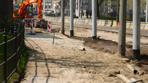 Chantier de voirie dans une rue montrant une pelleteuse orange sur un terrain sablonneux en cours d'excavation. &Agrave; gauche, une grille noire en fer forg&eacute; d&eacute;limite le chantier. En arri&egrave;re-plan, on aper&ccedil;oit des rails de tramway, des poteaux &eacute;lectriques et les fa&ccedil;ades &eacute;troites sous un ciel clair.