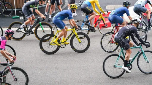 Vue en plong&eacute;e d'un groupe de cyclistes en pleine comp&eacute;tition sur un circuit urbain. Les coureurs, &eacute;quip&eacute;s de casques et de tenues de cyclisme aux couleurs vives (bleu, jaune, noir, rose), man&oelig;uvrent leurs v&eacute;los de course sur une route en asphalte.
