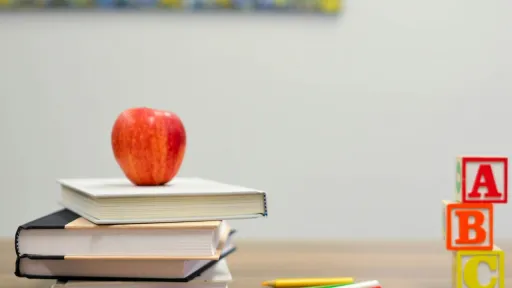 Une pile de trois livres surmont&eacute;e d'une pomme rouge, &agrave; c&ocirc;t&eacute; de quelques crayons de couleur et de trois cubes alphab&eacute;tiques empil&eacute;s affichant les lettres A, B et C sur un bureau en bois.