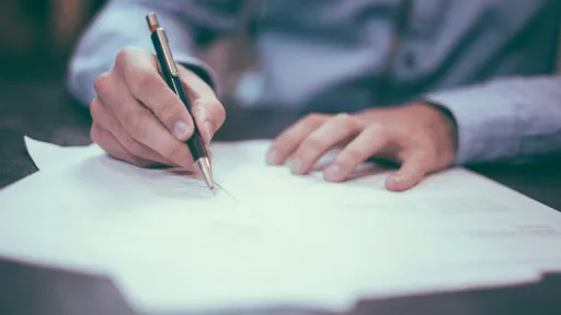 Gros plan sur les mains d'une personne portant une chemise bleue, tenant un porte-mine et &eacute;crivant ou signant sur une pile de documents blancs pos&eacute;s sur une table sombre.
