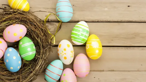 Plusieurs &oelig;ufs de P&acirc;ques aux couleurs pastel (rose, bleu, jaune, vert) d&eacute;cor&eacute;s de rayures et de pois, dispos&eacute;s &agrave; l'int&eacute;rieur et autour d'un nid de brindilles sur un fond de planches en bois clair.