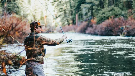 Une personne en cuissardes et veste marron se tient au milieu d'une rivi&egrave;re et pratique la p&ecirc;che &agrave; la mouche. Sa canne est lev&eacute;e et la ligne dessine une courbe &eacute;l&eacute;gante dans les airs. La sc&egrave;ne se d&eacute;roule dans un cadre naturel paisible, entour&eacute; d'une for&ecirc;t dense aux couleurs automnales.