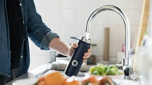 Gros plan d'une personne remplissant une gourde bleu fonc&eacute; au jet d'eau d'un robinet de cuisine chrom&eacute;. En arri&egrave;re-plan, on aper&ccedil;oit un mur carrel&eacute; blanc et au premier plan, un bol de fruits et de l&eacute;gumes flous.