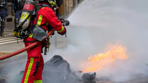 Un pompier en tenue rouge et jaune, portant un casque argent&eacute; et un appareil respiratoire, projette un puissant jet d'eau sur un feu au milieu d'une rue goudronn&eacute;e. De la fum&eacute;e blanche se d&eacute;gage du foyer.
