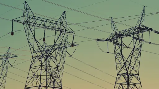 Silhouette de plusieurs pyl&ocirc;nes &eacute;lectriques et d'un r&eacute;seau de c&acirc;bles &agrave; haute tension se d&eacute;coupant sur un ciel au d&eacute;grad&eacute; de couleurs bleu-vert et orange doux.