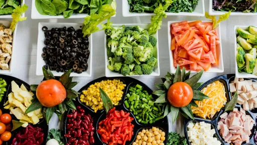 Vue a&eacute;rienne d'un buffet de bar &agrave; salade riche en couleurs, pr&eacute;sentant des bols de l&eacute;gumes verts (&eacute;pinards, chou fris&eacute;), des crudit&eacute;s (tomates, brocolis, carottes, olives), des l&eacute;gumineuses (pois chiches, haricots rouges) et des prot&eacute;ines d&eacute;coup&eacute;es, le tout organis&eacute; de mani&egrave;re sym&eacute;trique sur un fond blanc.