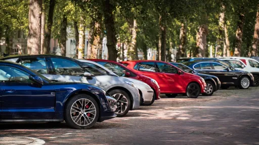 Une rang&eacute;e de voitures gar&eacute;es en &eacute;pi le long d'une route pav&eacute;e, &agrave; l'ombre de grands arbres feuillus. Au premier plan, on distingue nettement le profil d'une voiture &eacute;lectrique bleu marine. En arri&egrave;re-plan, plusieurs autres v&eacute;hicules de diff&eacute;rentes couleurs (gris, rouge, noir et blanc) sont align&eacute;s sous la lumi&egrave;re tamis&eacute;e du soleil filtrant &agrave; travers le feuillage.